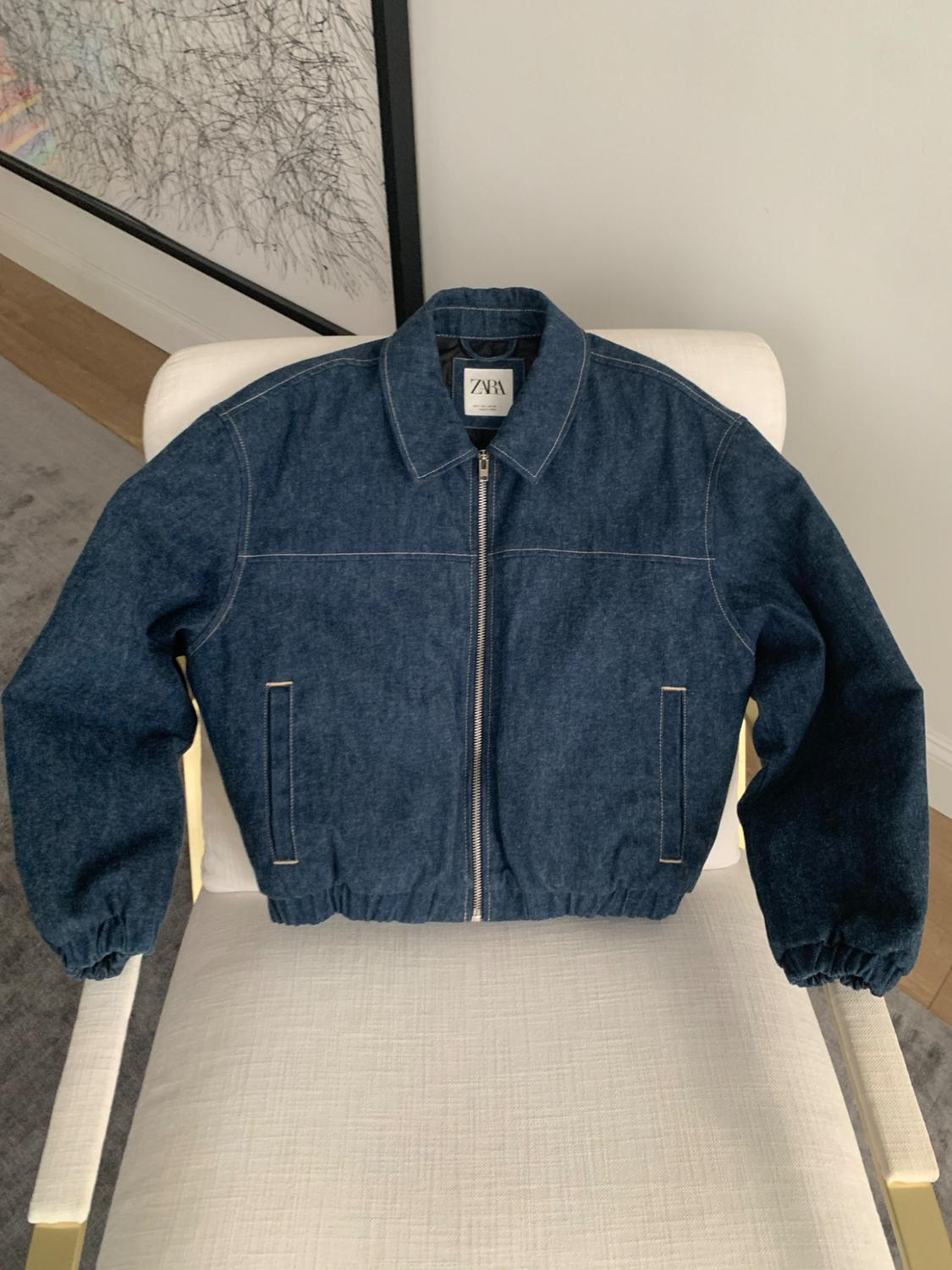 Blue denim jacket with a zipper, elastic hem, and ZARA label, resting on a white armchair. An abstract painting is visible in the background.