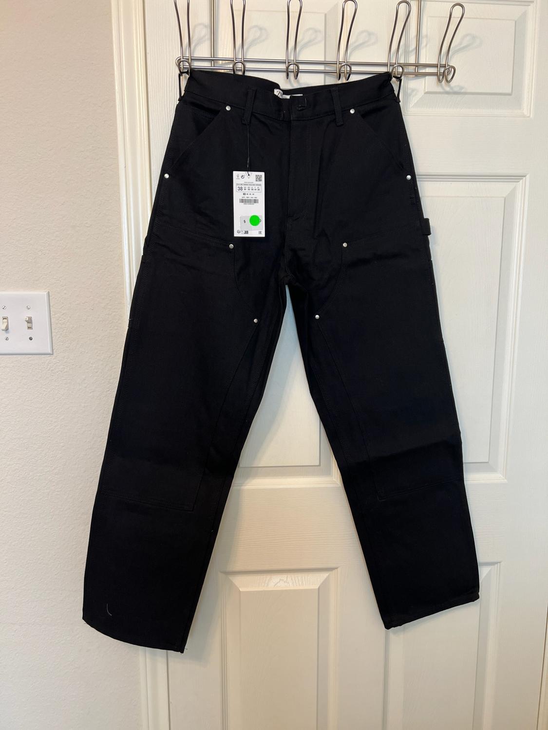 Black trousers with silver studs and a label, hanging from a metal clothes rail over a white door.