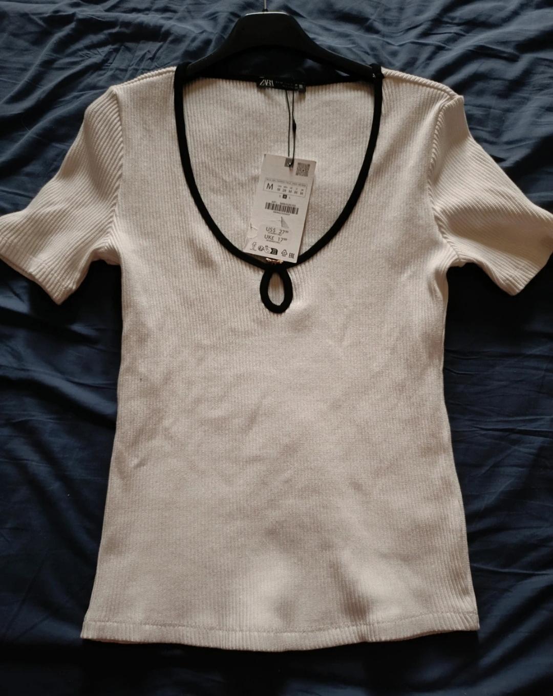 Beige short-sleeved T-shirt with ribbed texture, V-neck with black trim, hanging on a hanger over blue fabric. Label visible.