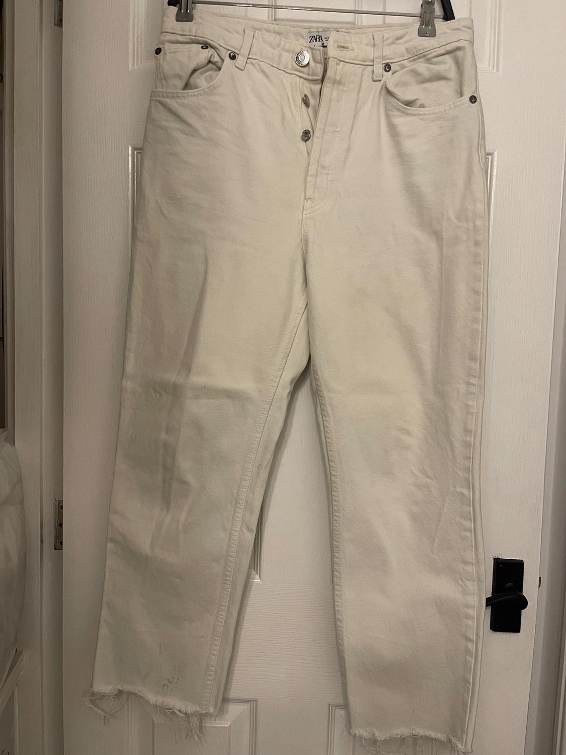 Beige denim trousers hanging on a white door, with visible buttons and a frayed hem at the bottom.