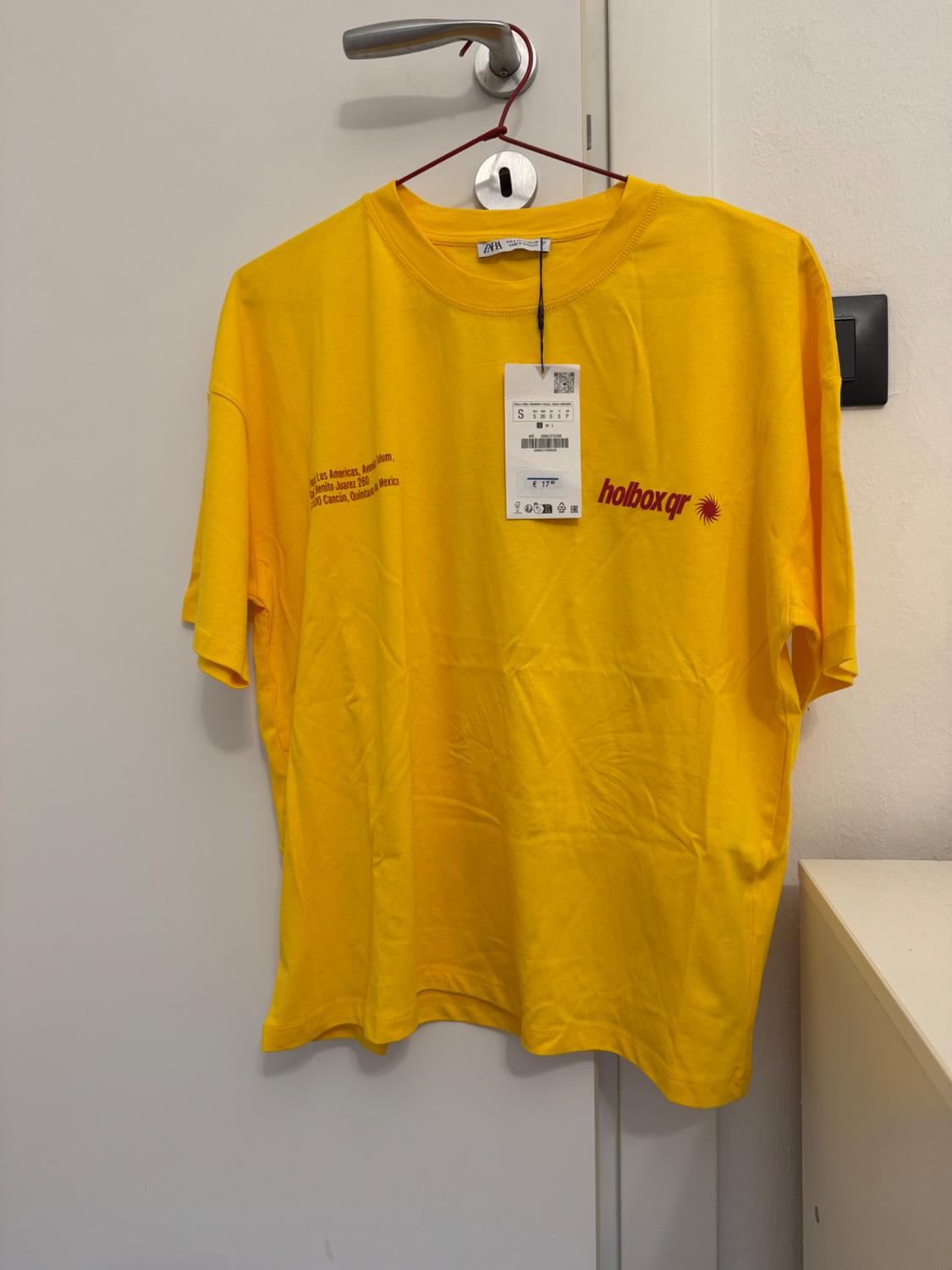 Yellow t-shirt hanging on a door, with "holbox qr" text and address on the chest. Price tag visible.