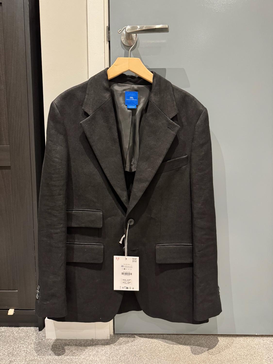 Black blazer hanging on a wooden hanger on a grey door. Blue inside ZARA label and white hanging tag with the text 48.