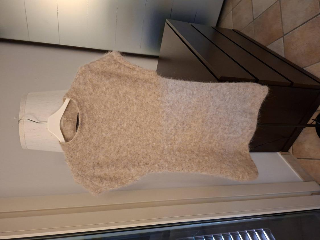 Beige knitted jumper hanging on a white hanger against a background of wooden furniture and floor tiles.