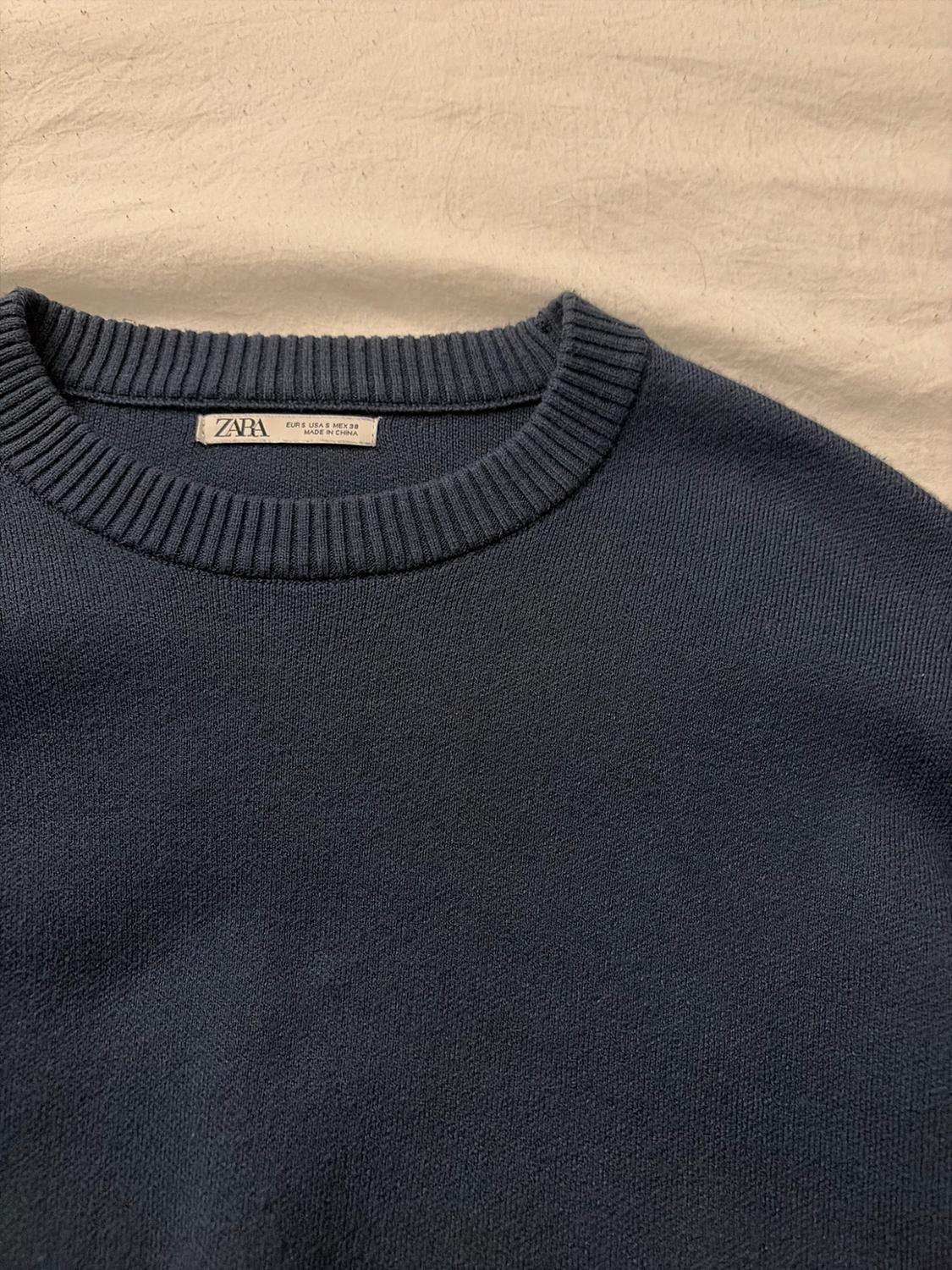 Close-up of a navy blue knit sweater on white fabric. The label at the neckline reads "ZARA EUR S USA S MEX 38 MADE IN CHINA".