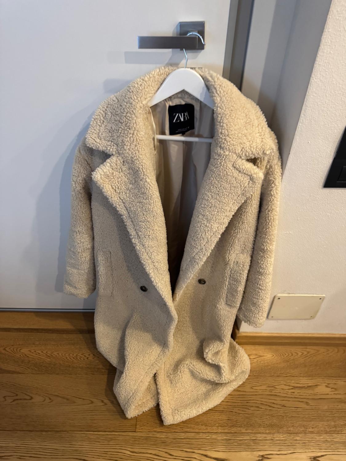 Beige wool coat hanging on a white hanger over a door. Visible label with the text "ZARA". Wooden floor.