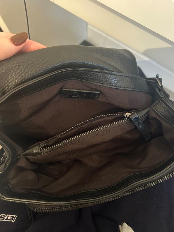 Black leather handbag, open, showing the brown interior with a zipped pocket. Hand with brown painted nails holding the handbag.