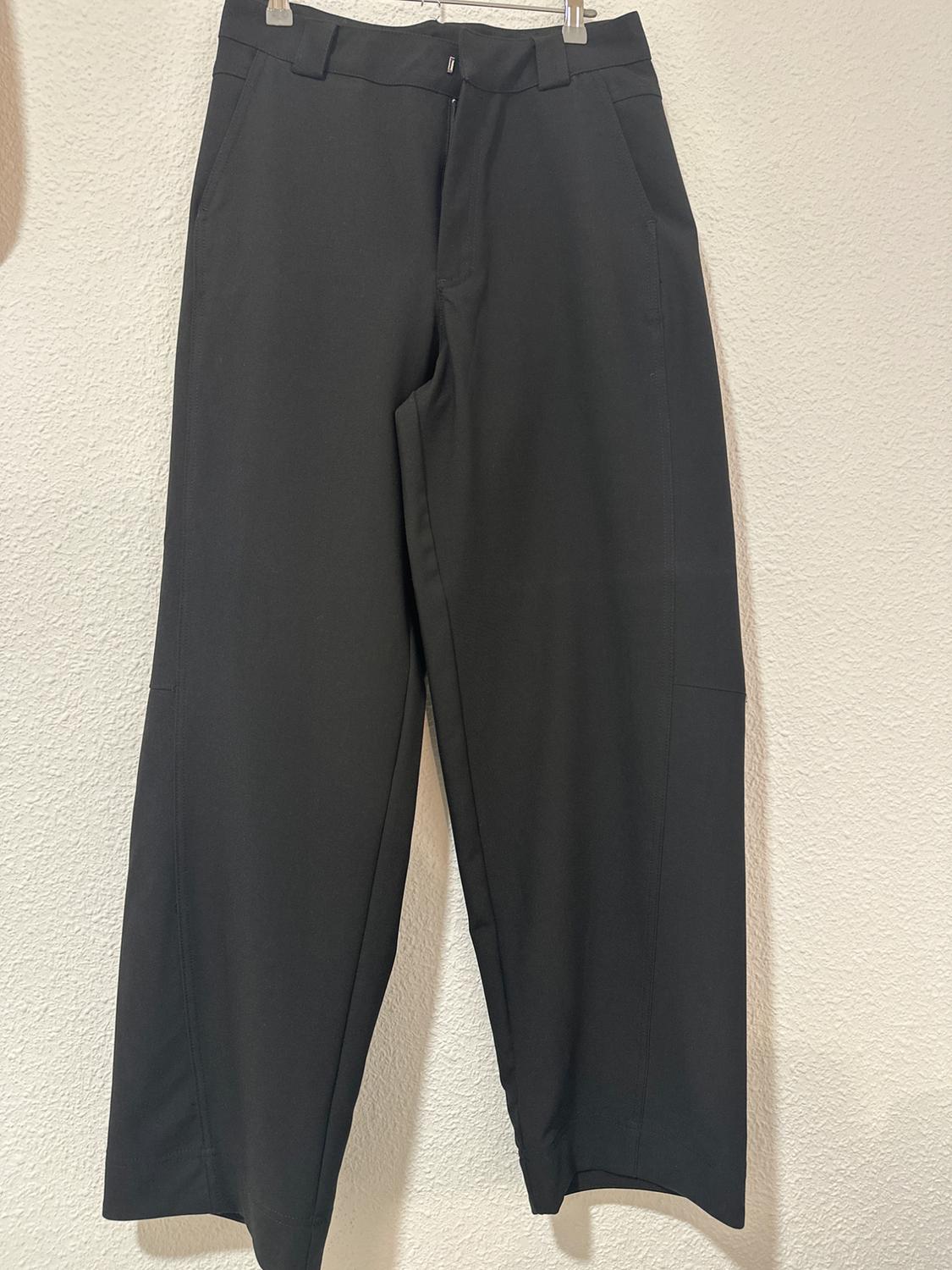 Black trousers hanging on a hanger against a white wall. Simple design with side pockets and belt loops.