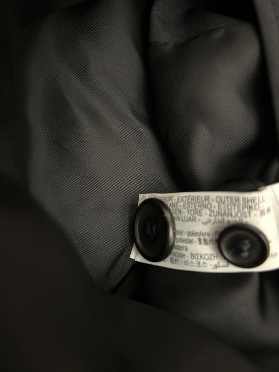 Care label on black fabric with visible black buttons. Text in various languages, including "EXTERIOR" and "OUTER SHELL".