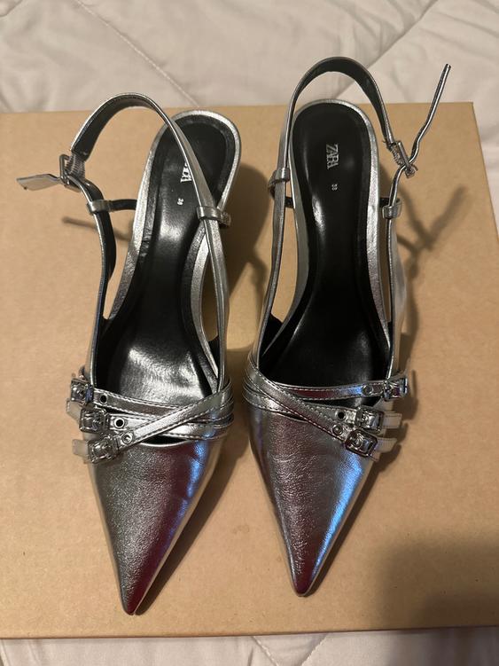 Silver high-heeled shoes with a pointed toe and adjustable straps, on a brown box. Visible text: "ZARA".