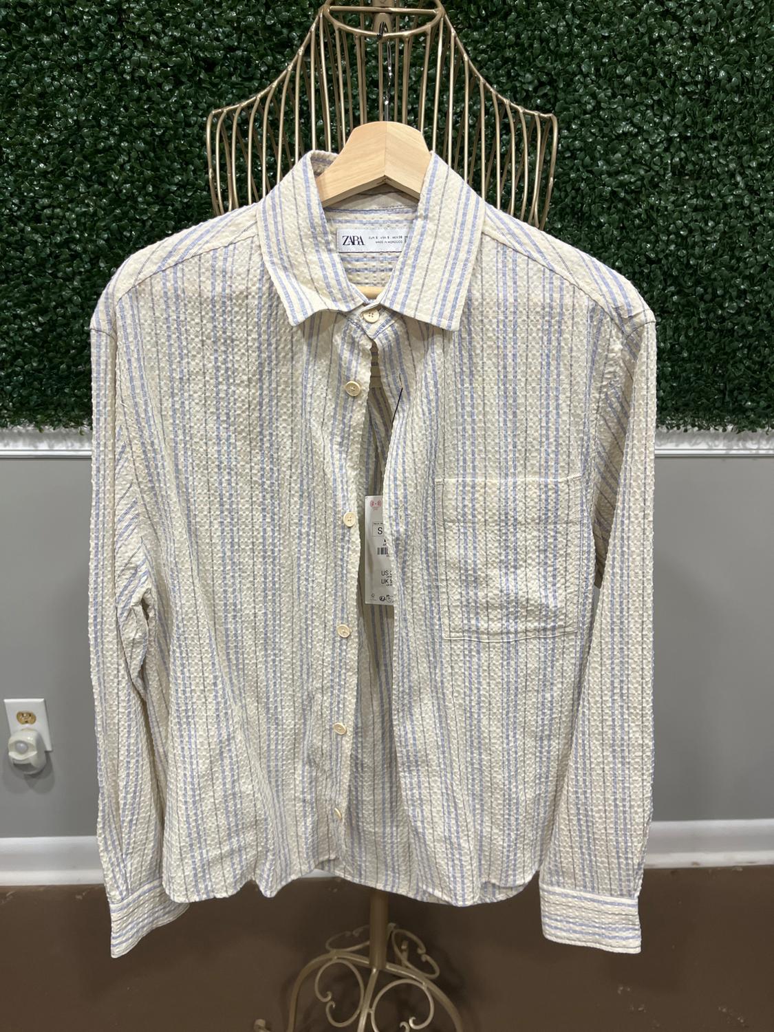 Vertical striped shirt in beige and light blue tones hanging on a wire mannequin, green wall background. Visible tag.