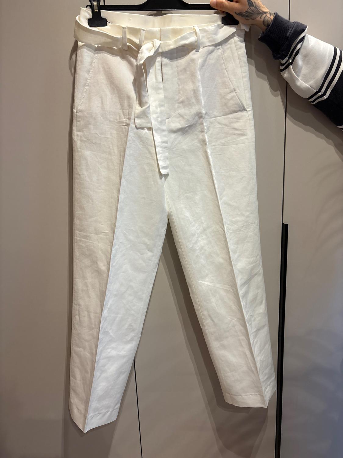 White trousers are hanging on a hanger, accompanied by a matching belt. A tattooed hand is holding the hanger. The background is neutral.