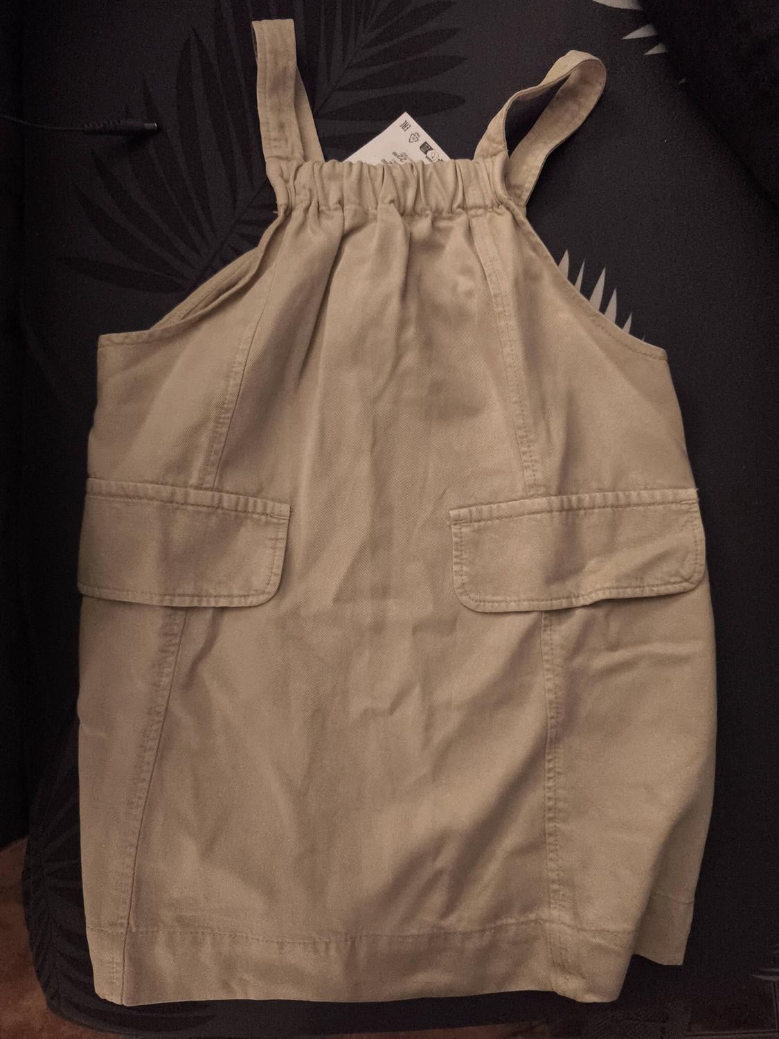 Beige dress with straps and front pockets, on a dark background with a leaf design. Visible label at the top.