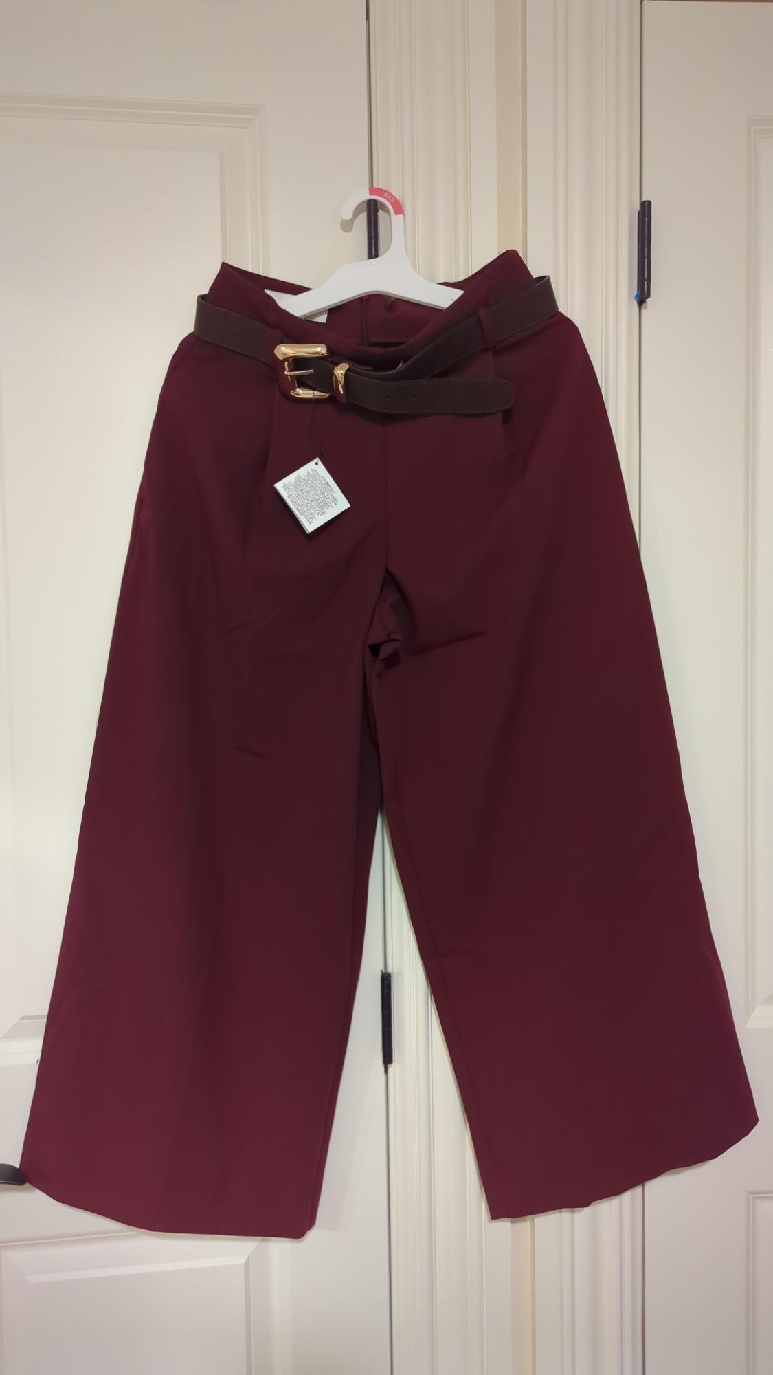 Burgundy trousers hanging on a white hanger, with a brown belt and visible label, against a background of white doors.