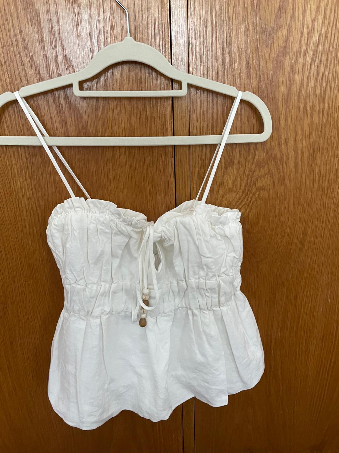 White sleeveless top with spaghetti straps and adjustable drawstring, hanging on a beige hanger against a wooden background.