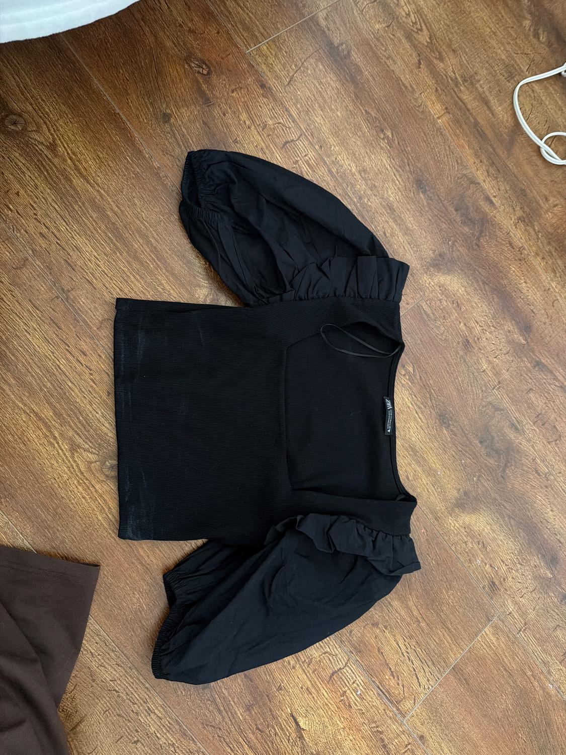 Black long-sleeved puffed blouse on a wooden floor. Part of a brown garment visible in the bottom left corner.