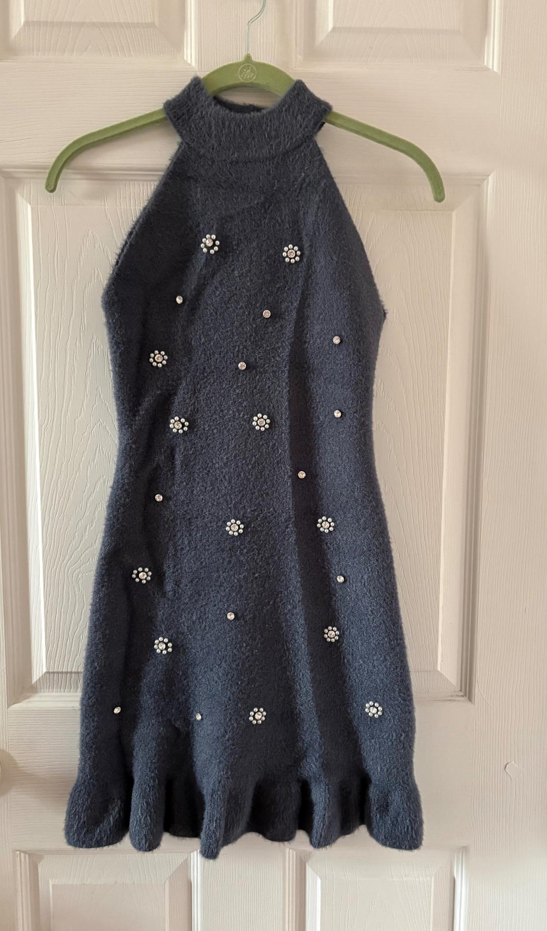 Dark blue, sleeveless, high-neck dress, decorated with pearl and floral appliques, hanging on a green hanger in front of a white door.