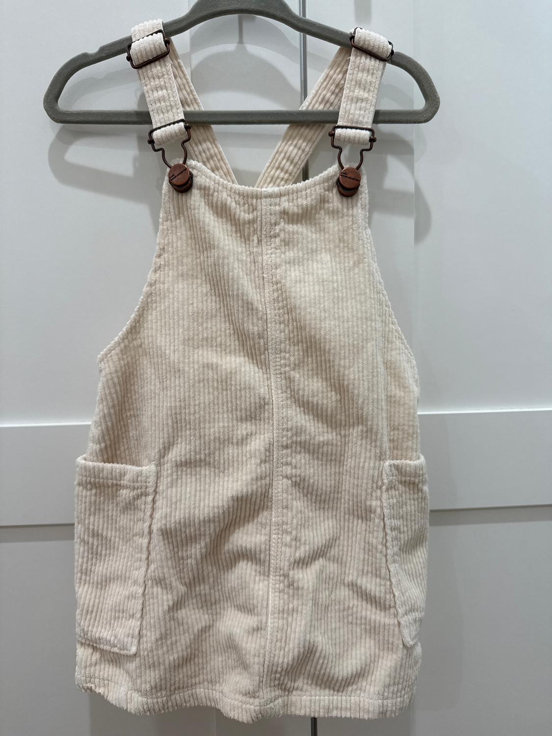 Beige corduroy dungarees hanging on a grey hanger, with adjustable straps and front pockets, on a white background.