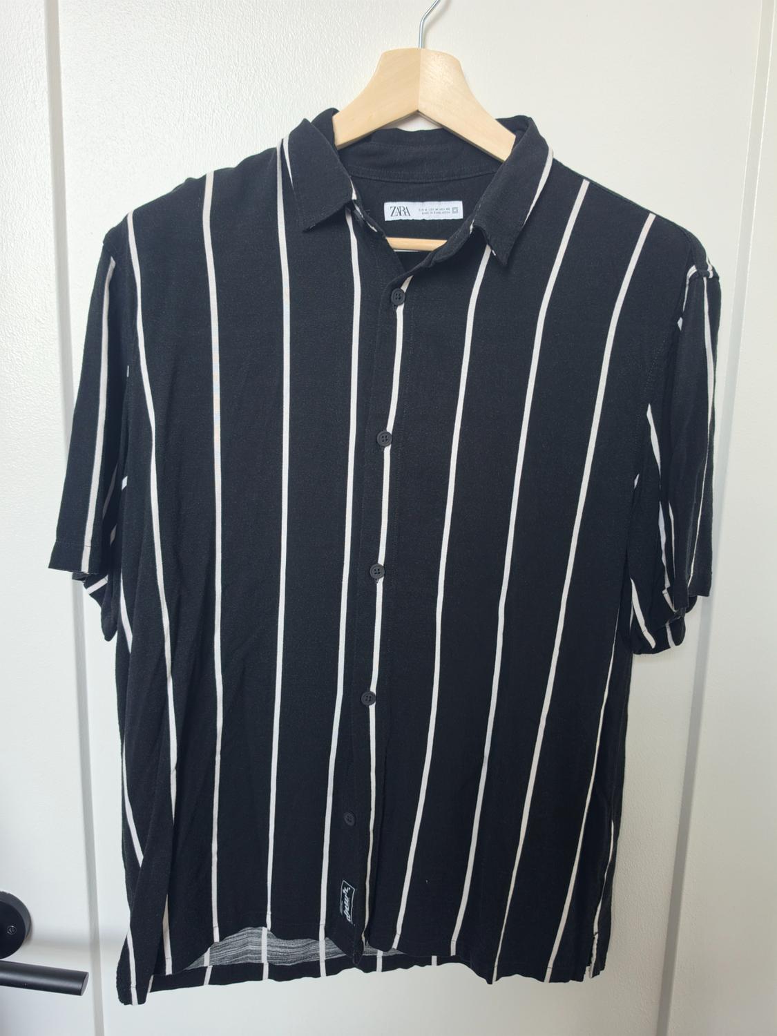 Black short-sleeved shirt with white vertical stripes hanging on a wooden hanger against a white door. ZARA text.