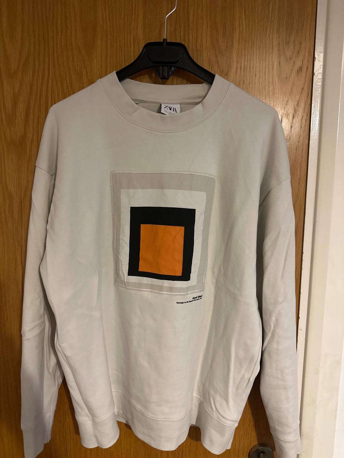 Front view of white sweatshirt with square geometric print in black, orange and small text underneath.