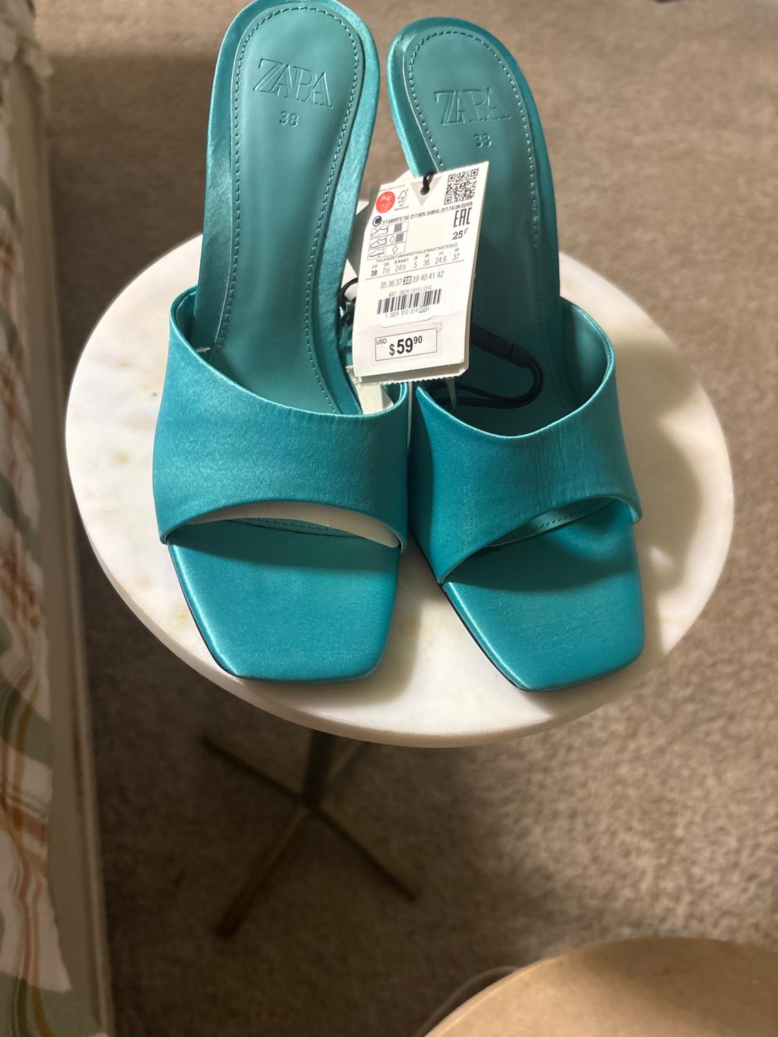 Turquoise low-heeled sandals with a $59.90 price tag, placed on a round marble table.