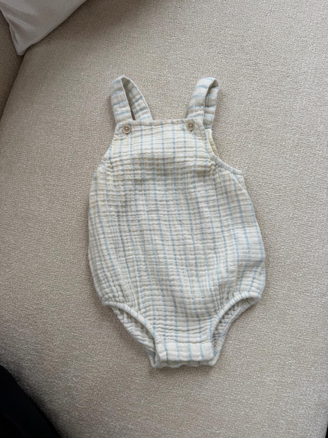 Cream-colored muslin fabric baby romper with a print of thin vertical blue stripes, buttoned shoulder straps, and a diaper-style cut.