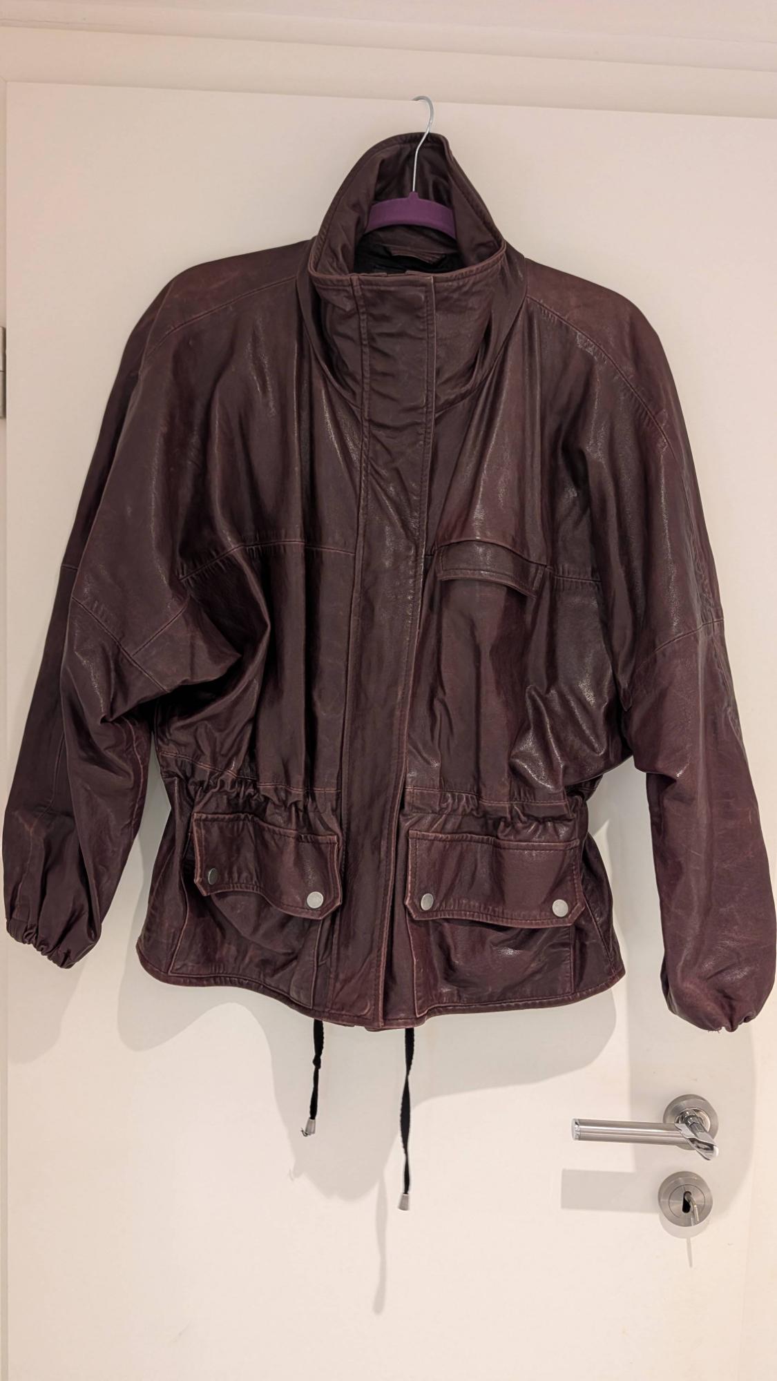 Dark brown leather jacket hanging on a white door. It features a high collar, elasticated waist, front pockets and drawstrings at the hem.