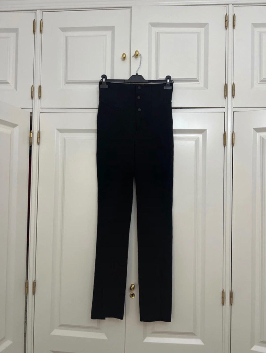 Black high-waisted trousers with front buttons hanging on a hanger in front of a white wardrobe with gold handles.