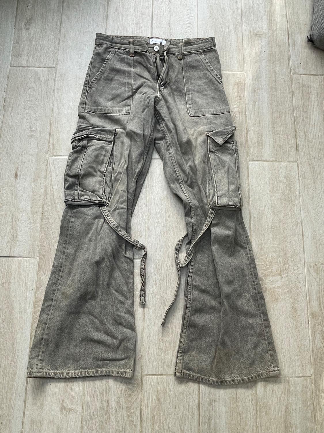 Grey cargo trousers with large pockets and decorative ribbons on the legs, on light wooden flooring.