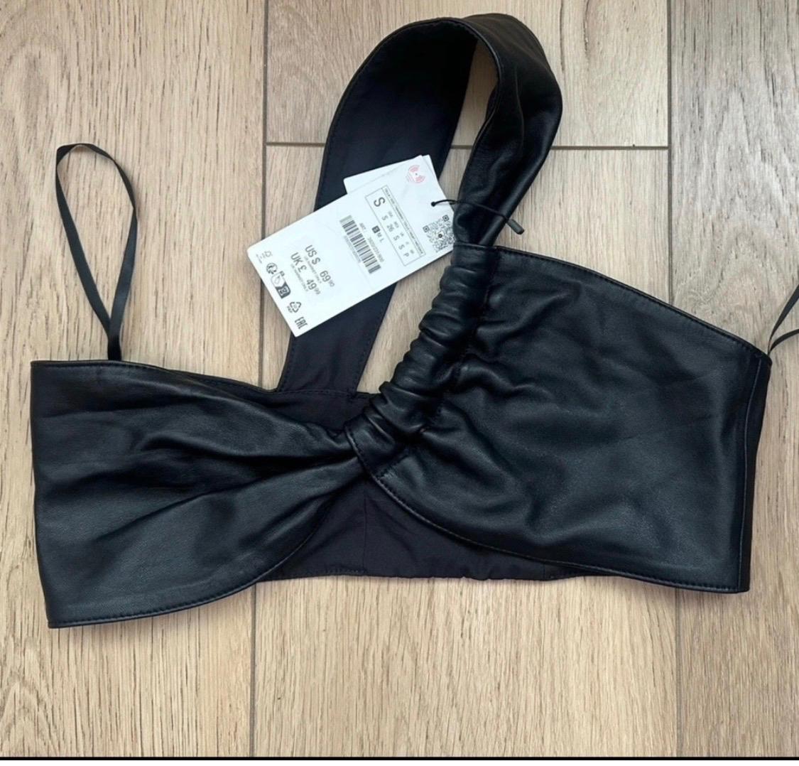 Asymmetrical black top with one wide gathered strap and one thin strap on a wooden floor. Label: S US $ 69.90 UK £ 49.99.