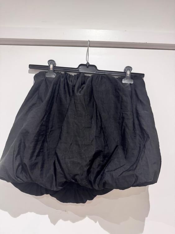 Puffball black skirt hanging on a hanger against a white wall. The fabric has a crinkled and voluminous finish.