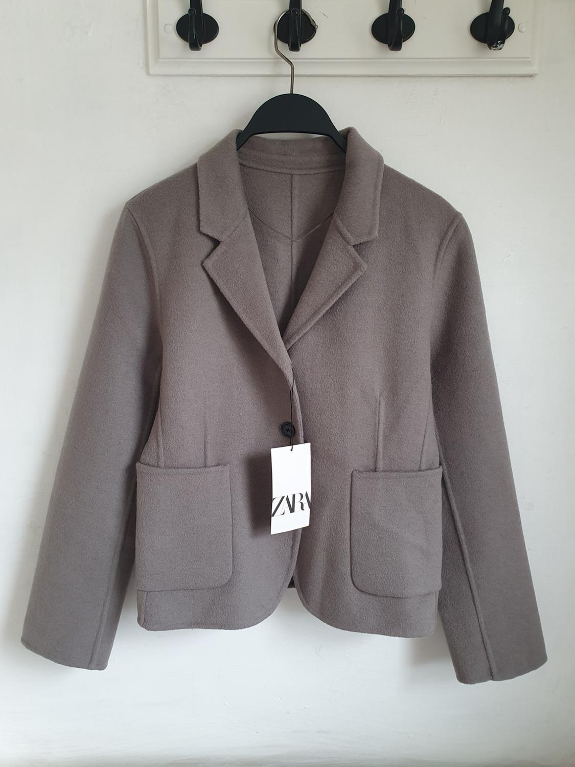 Grey jacket with lapels and large pockets, hanging on a black hanger. Visible label with the text "ZARA". White background.