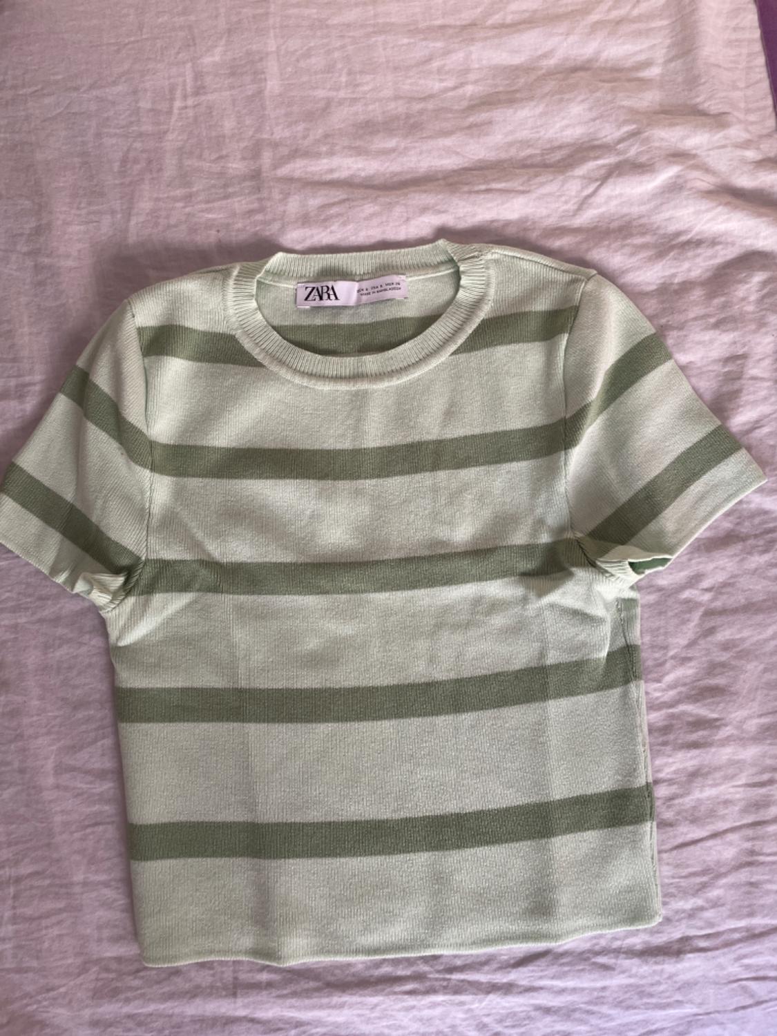 Green and white horizontal striped T-shirt on a wrinkled fabric background. Visible label with the text "ZARA".