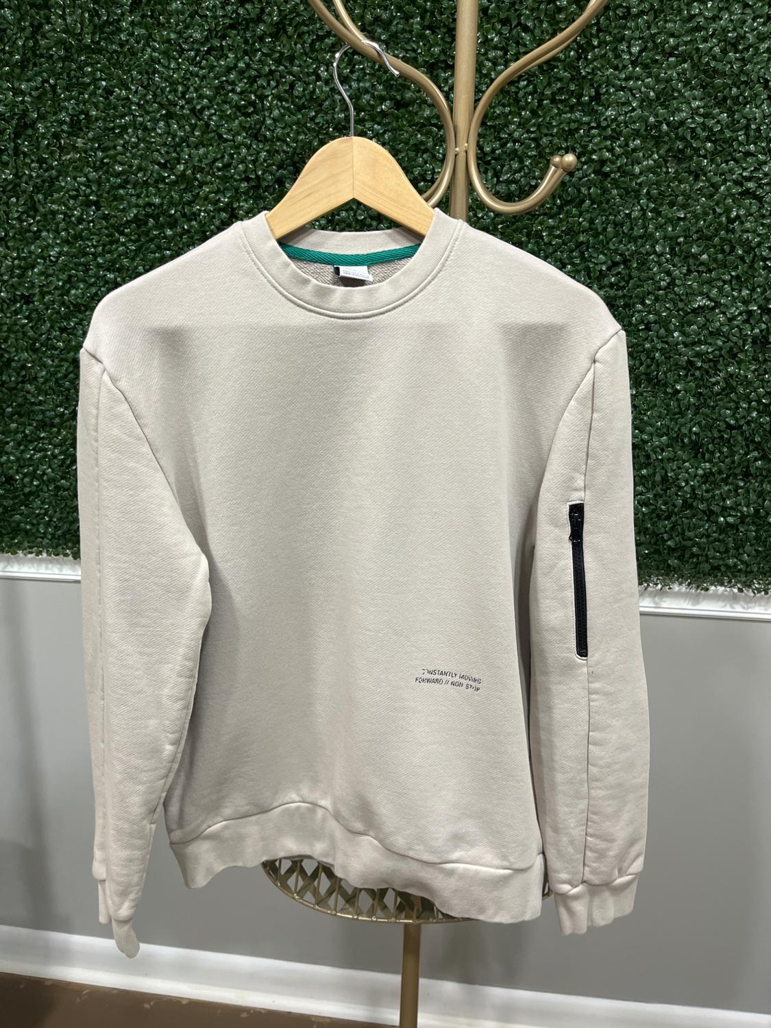 Beige sweatshirt hanging on a wooden hanger, with text "Instantly moving forward in new step" and zipper on the left sleeve. Green plant background.