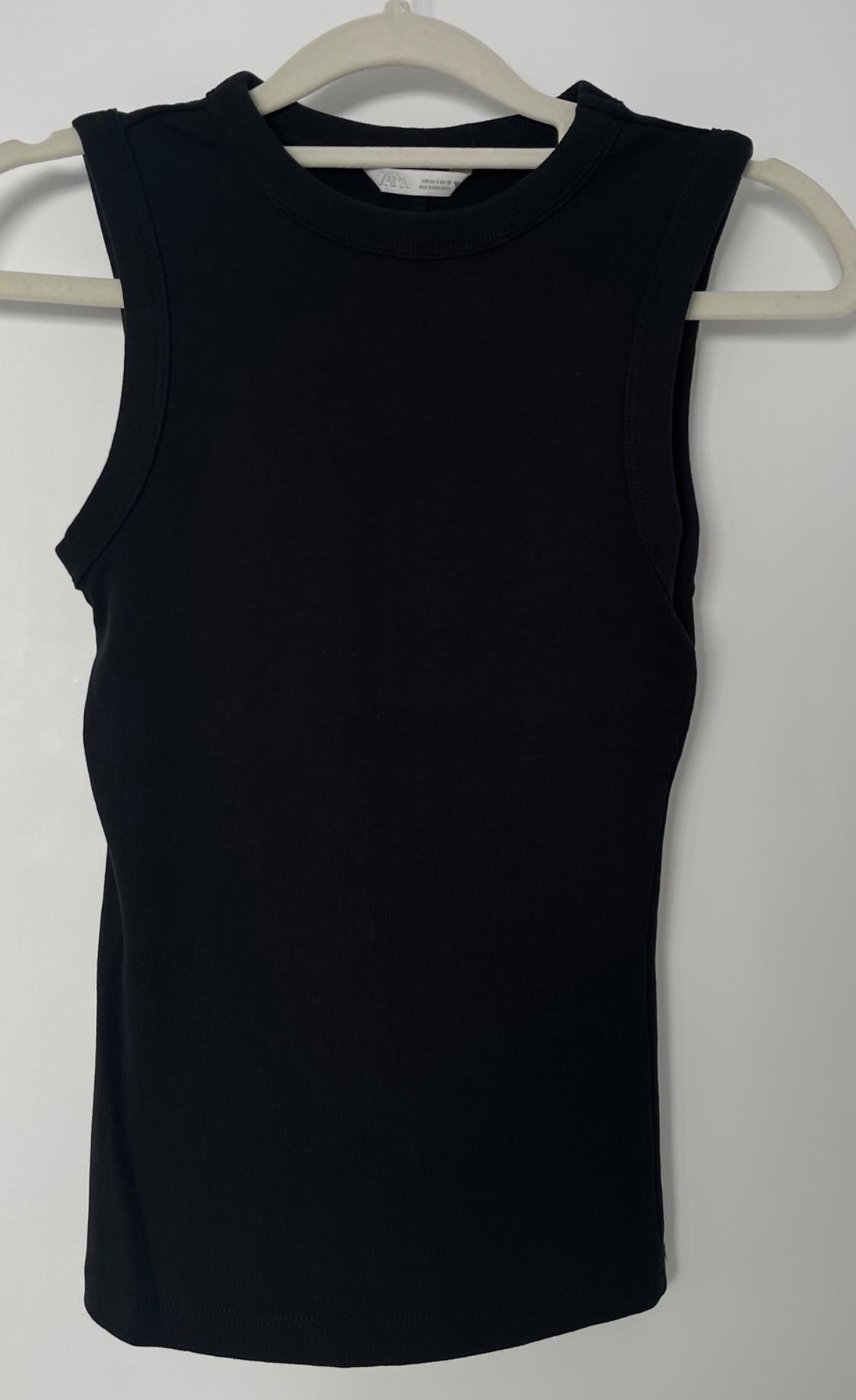 Black sleeveless top hanging on a white hanger against a light background. Visible label on the collar.