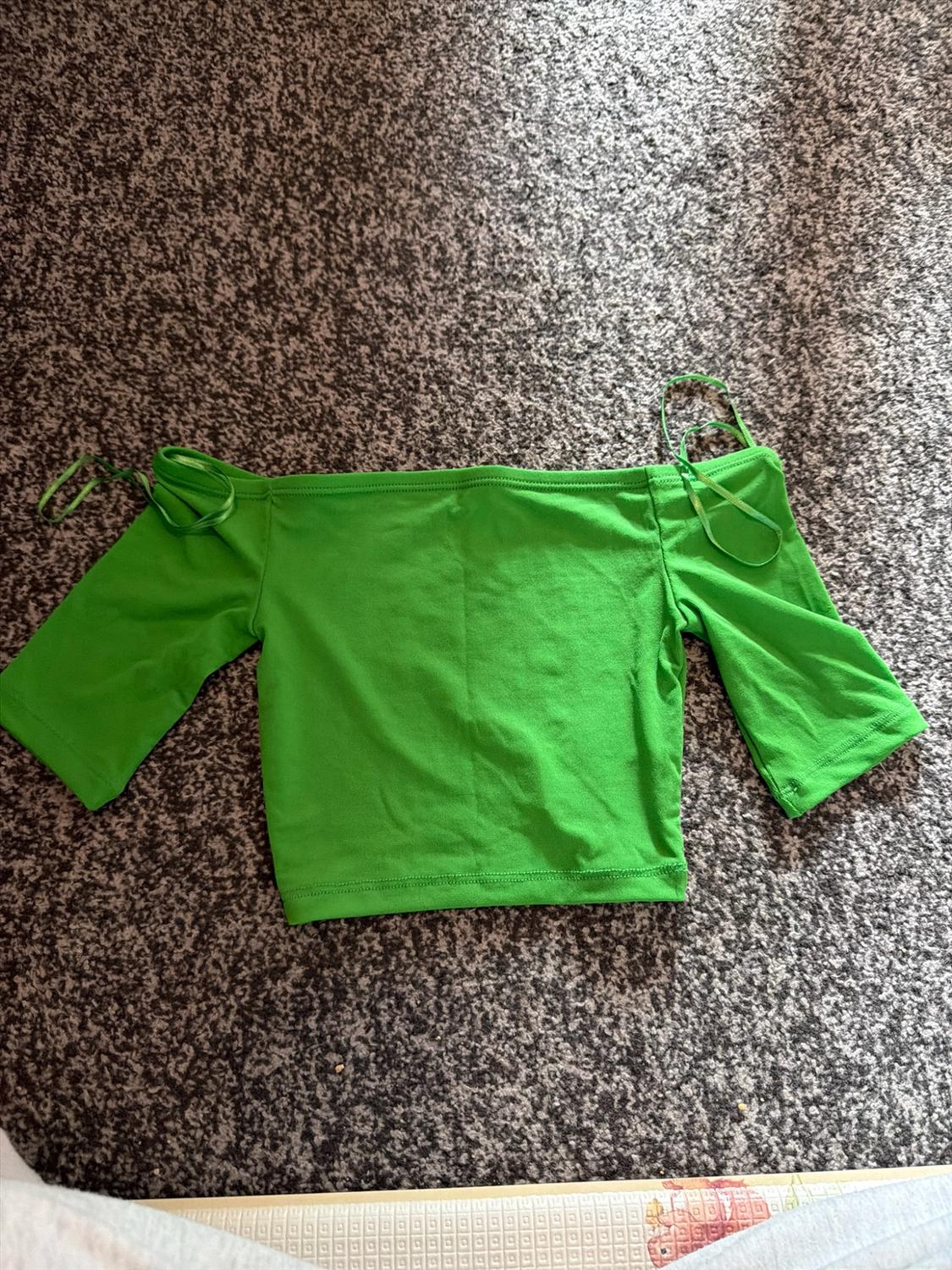 Cropped green off-the-shoulder top with short sleeves and thin straps, placed on a textured grey rug.