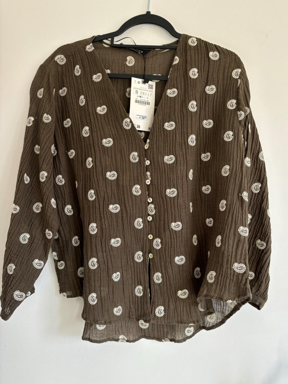 Brown blouse with white paisley print, front buttons and a €39.99 price tag hanging on a black hanger.