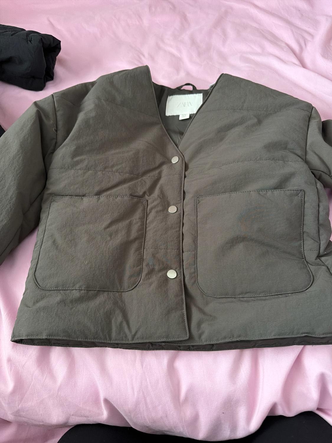 Olive green padded jacket with press studs, two front pockets and a ZARA label, set against a crinkled pink fabric.