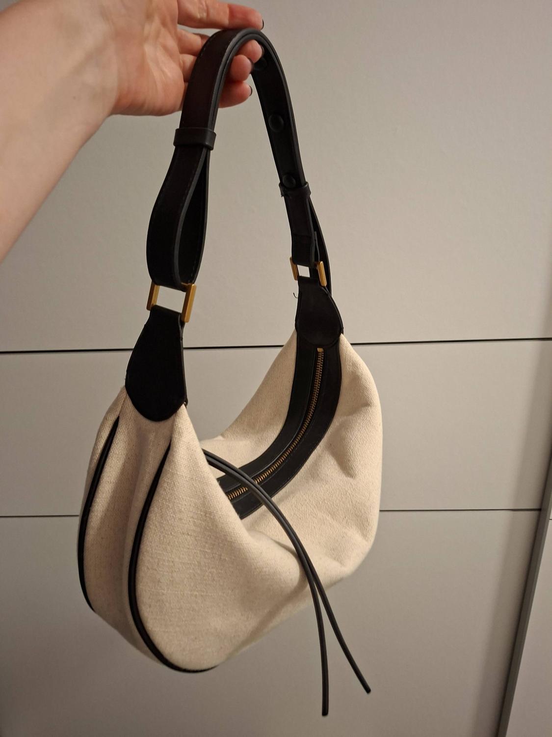 Person holding a light-coloured fabric shoulder bag with black trim and handle, and gold-tone hardware, against a panelled background.