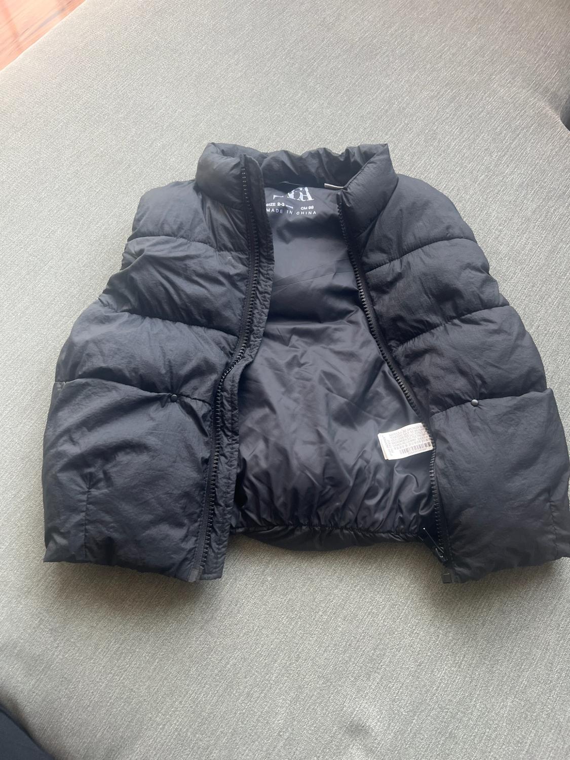 Black padded gilet with an open zip on a grey surface. Text: ZARA SIZE 2-3 YEARS CM 98 MADE IN CHINA.