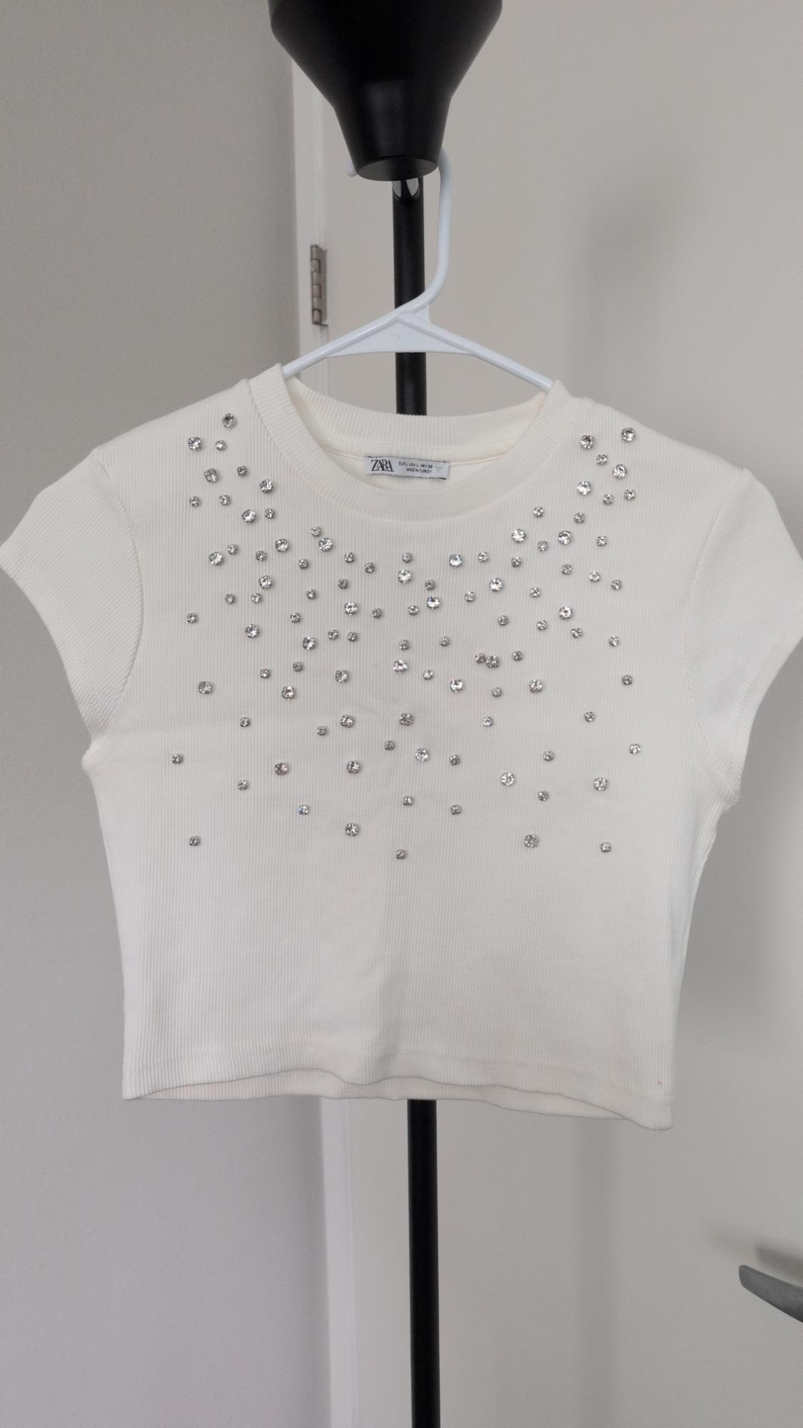 White ribbed short-sleeve cropped top with rhinestones, hanging on a hanger. Label with ZARA text.