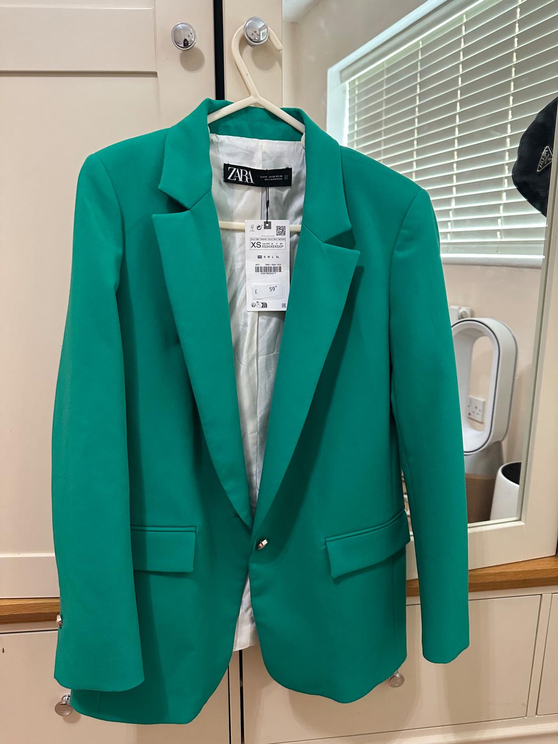 Green jacket hanging on a hanger with a visible Zara label, against a white wardrobe and mirror background.