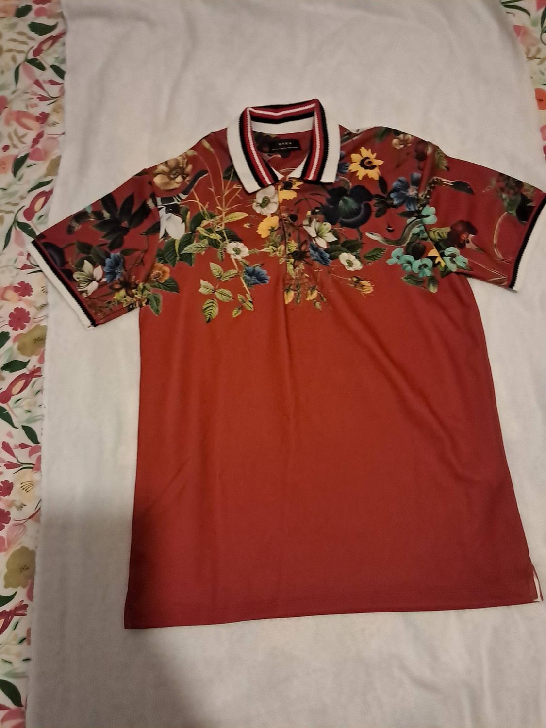 Red polo shirt with a floral print on the upper section and a striped white collar, on white fabric. ZARA text.