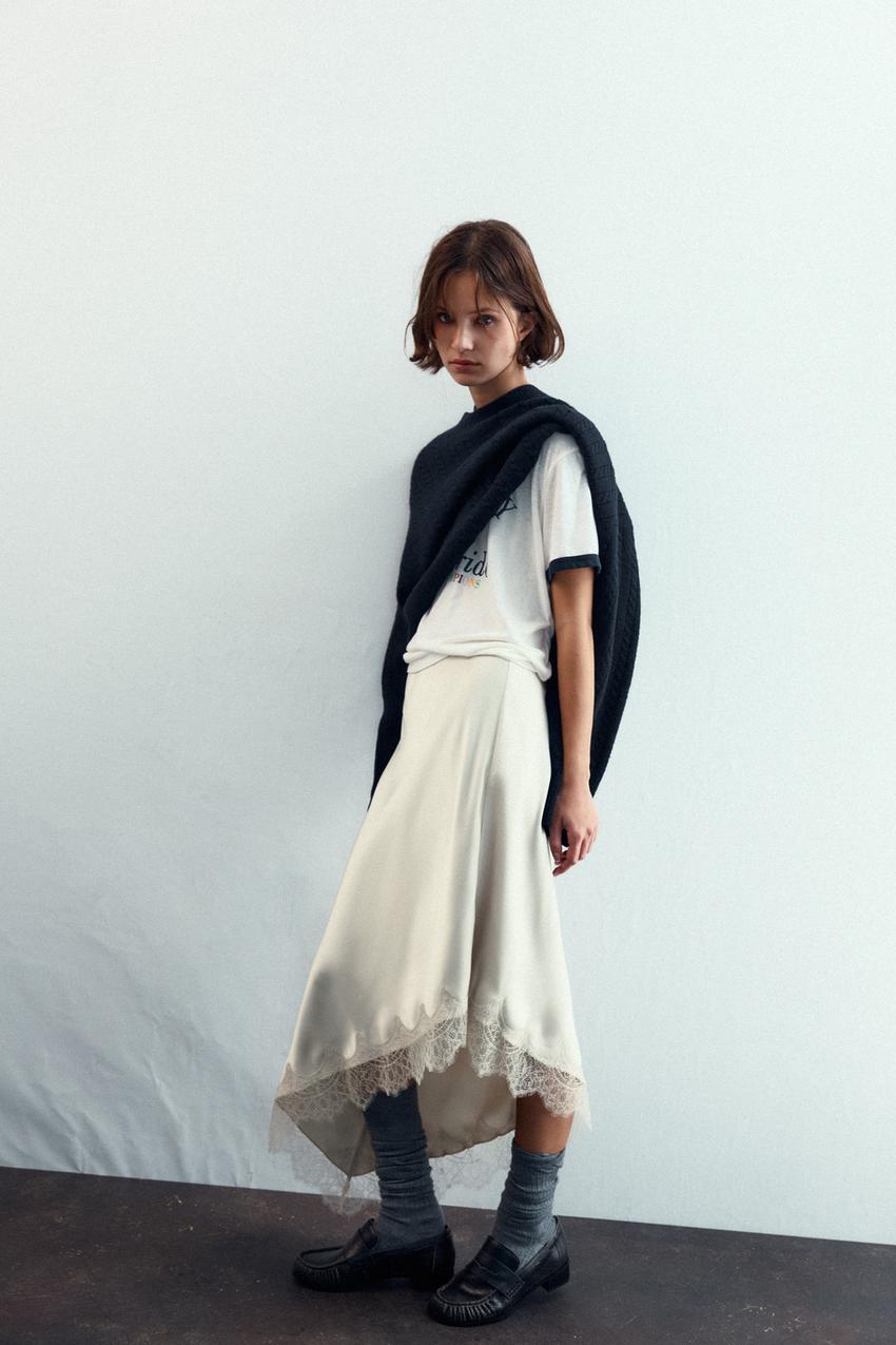 White t-shirt with text, beige lace skirt, black sweater over the shoulders, gray socks, and black loafers set.