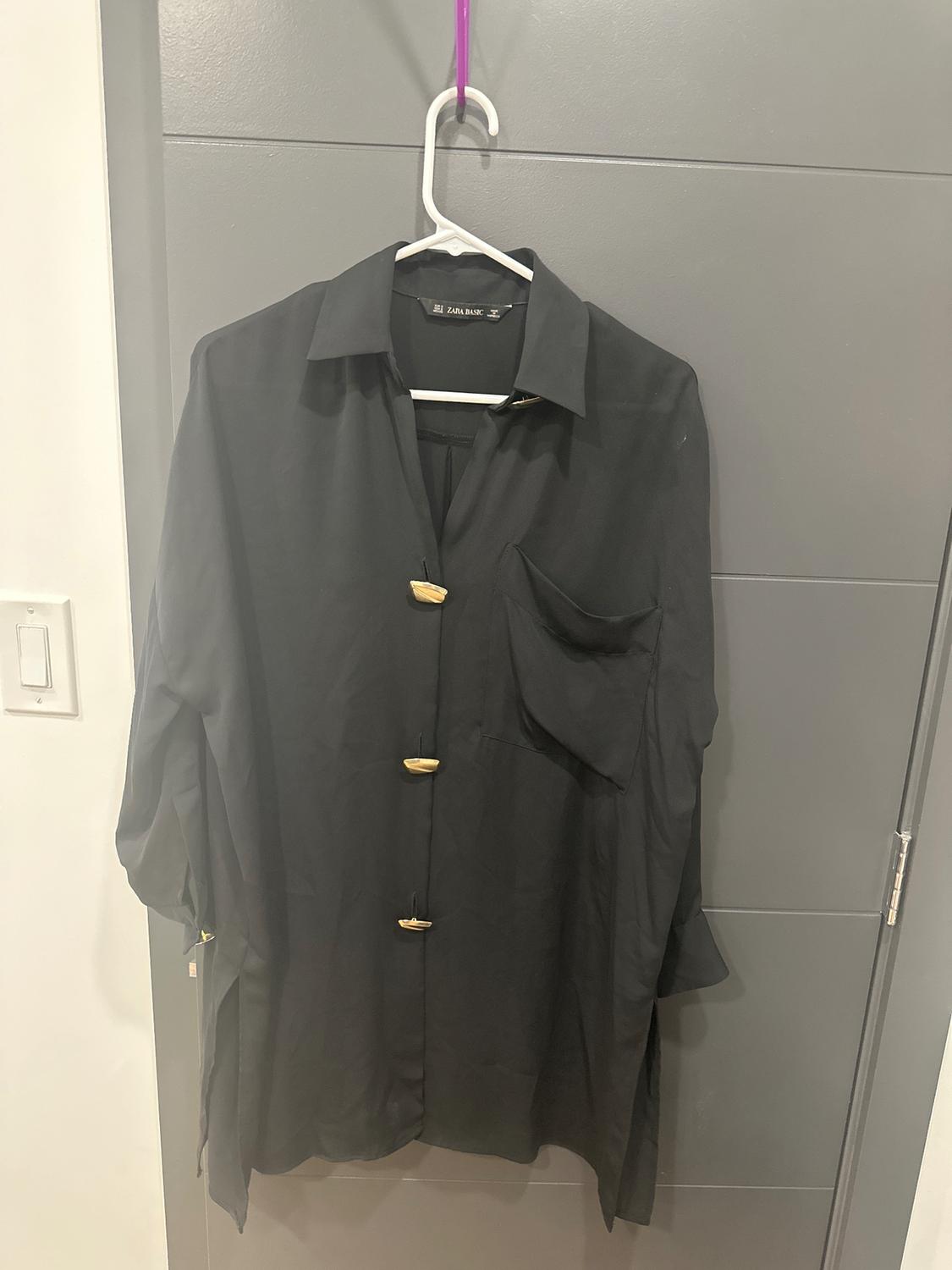 Black long-sleeved shirt hanging on a white hanger with golden buttons and a chest pocket. Gray door background.