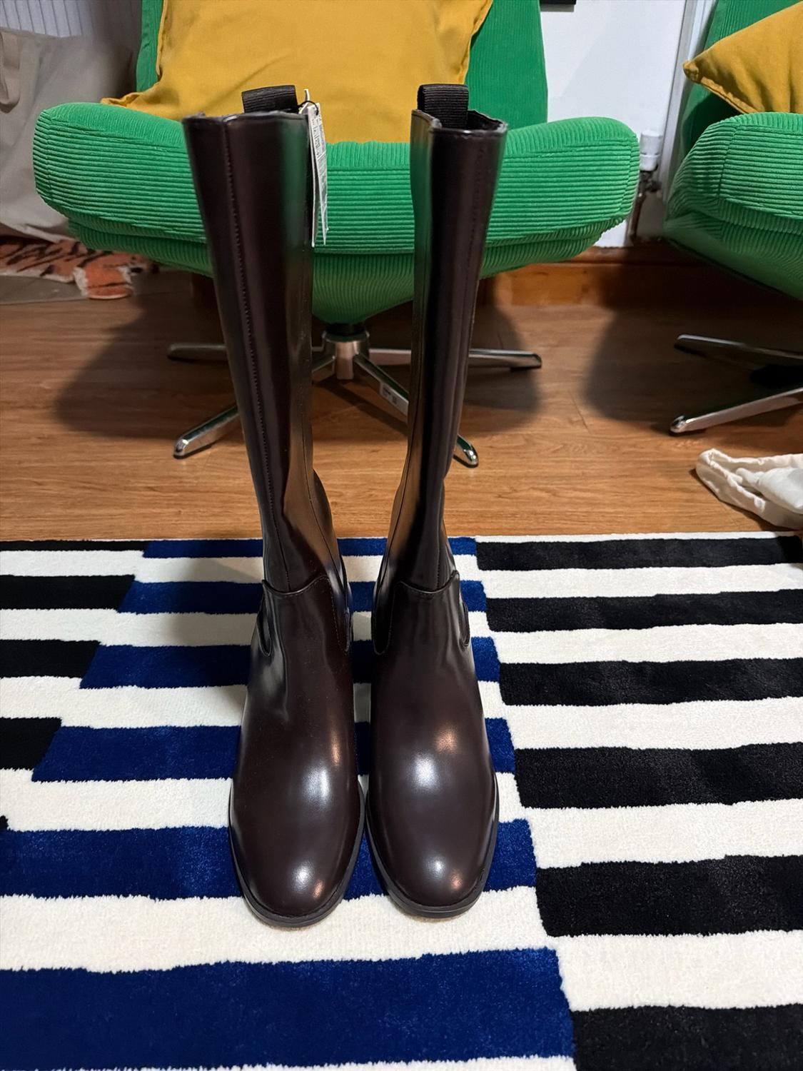 Dark brown knee-high boots on a white, black and blue striped rug. In the background, a wooden floor and green armchairs.