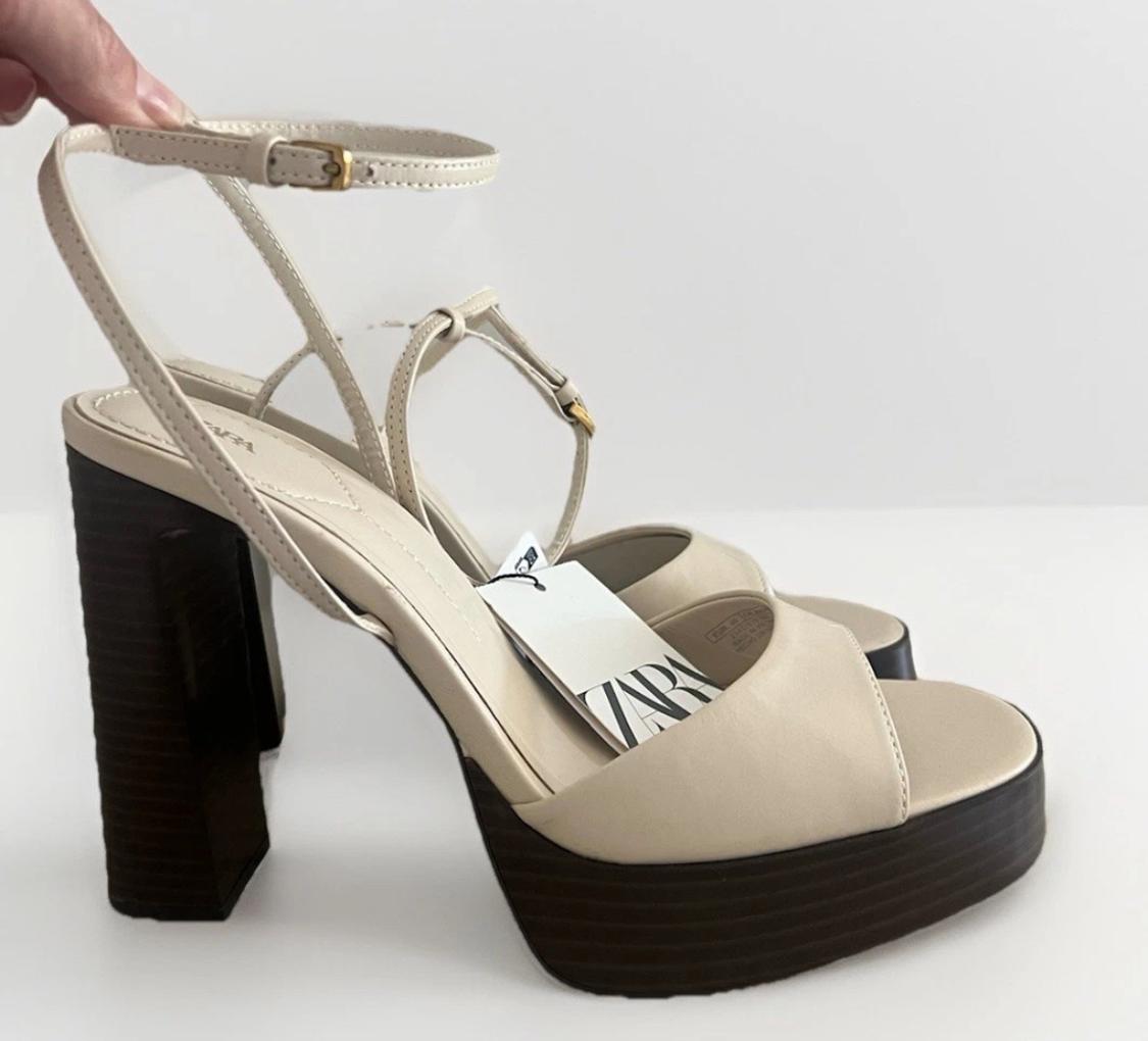 Here's the rephrased sentence, maintaining the same meaning and context, without using any of the forbidden terms: "A hand holds a pair of beige strappy sandals, with a brown heel and platform, against a white background. They have a label with the text ZARA."