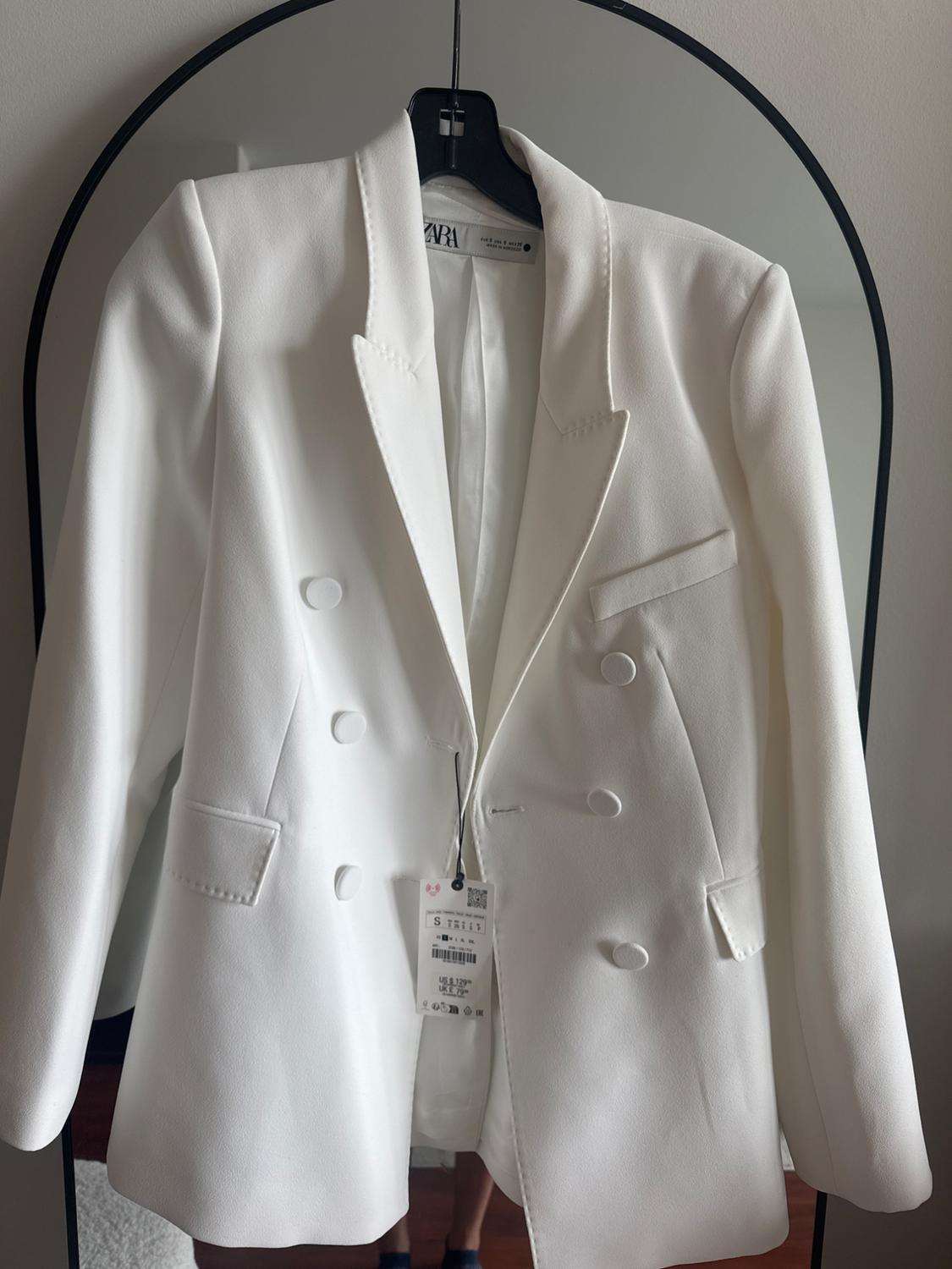 White double-breasted jacket hanging on a mirror. Visible label with "ZARA" text and size and price details.