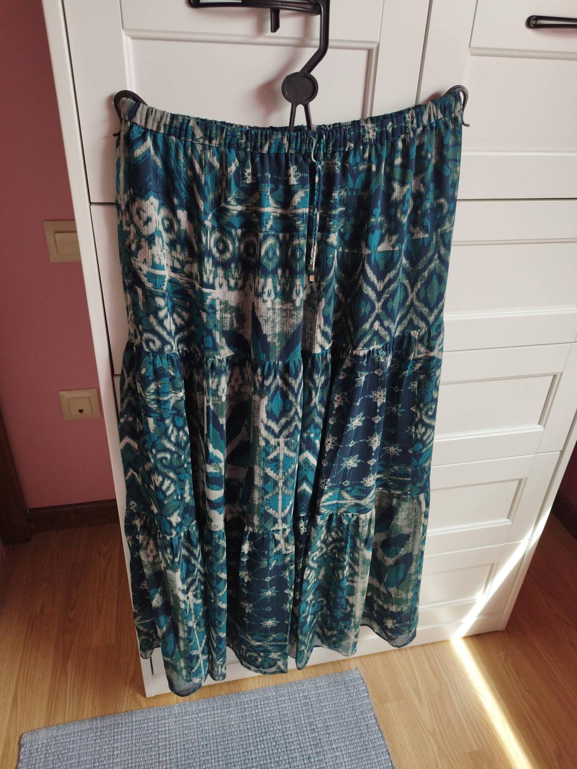 Long skirt with blue and green abstract print hanging from a hanger in front of a white cabinet. Wooden floor and natural light.