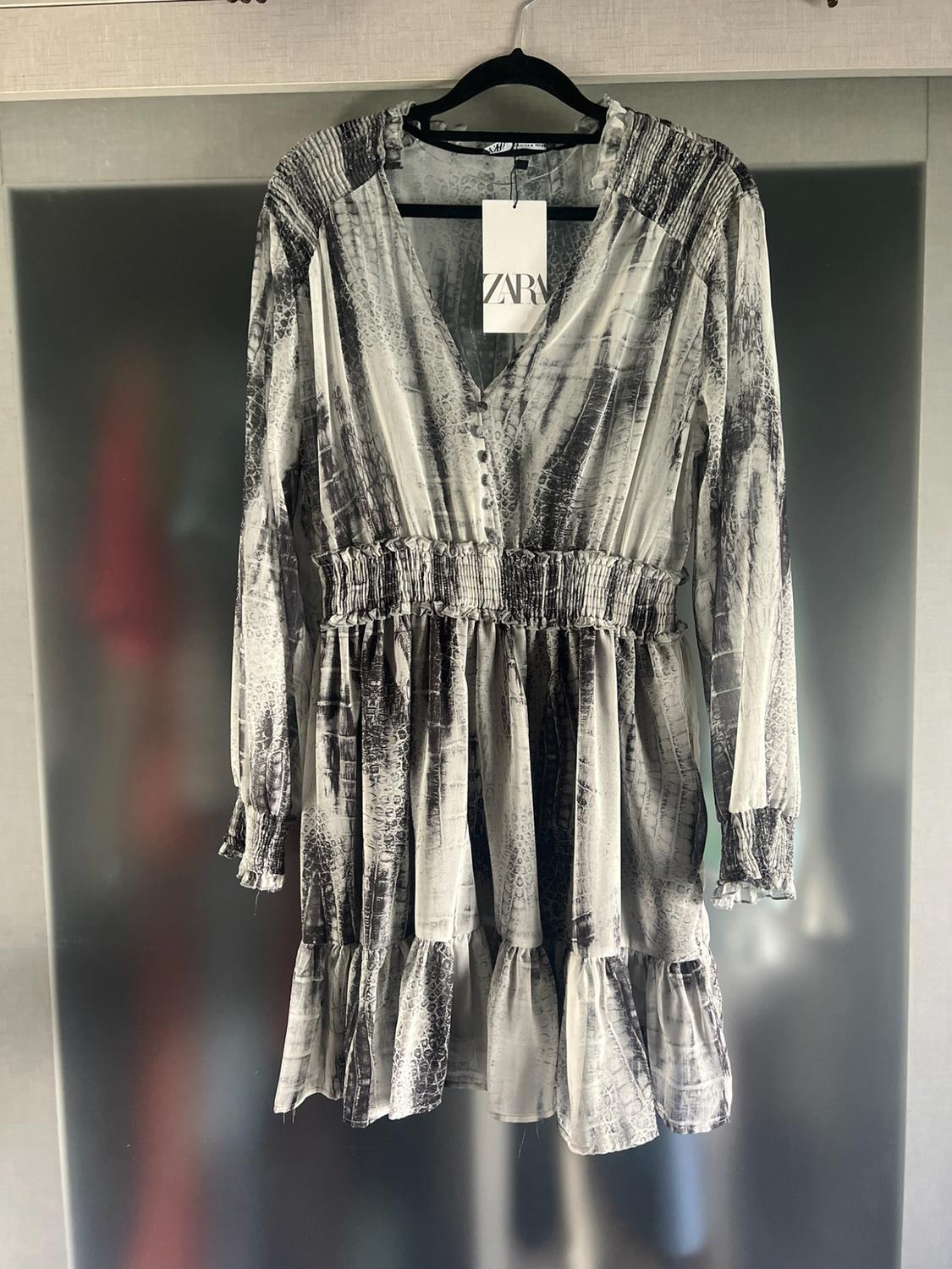 Short long-sleeved dress with a black-and-white print hanging on a hanger. It has an elasticated waist and a label bearing the text ZARA.
