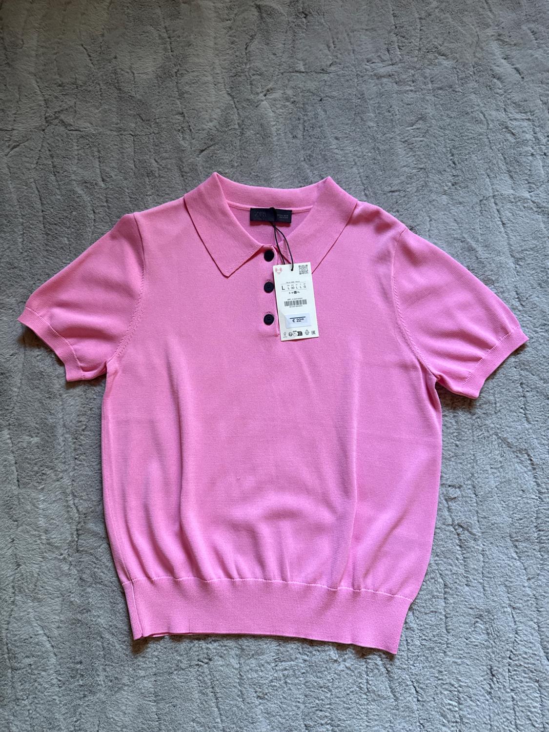 Pink polo shirt with a classic collar and black buttons on a grey background. Visible hang tag in the centre.